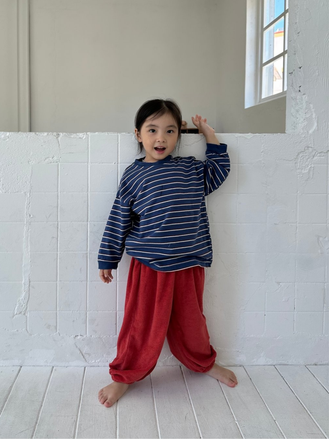 Blue | A young child stands barefoot indoors against a white tiled wall, wearing the PEPPER LONGSLEEVE TEE with blue stripes and red pants, one arm raised and mouth slightly open.