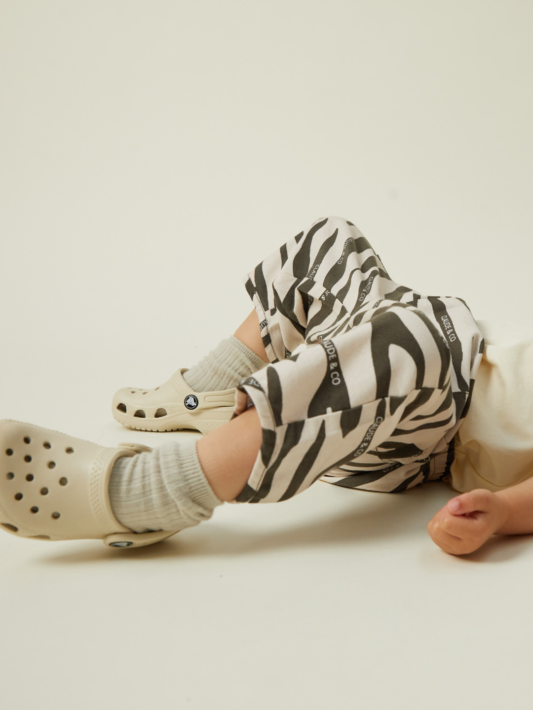 A child dressed in EMERSON ZEBRA TROUSER, cozy beige clogs, and gray socks lies comfortably on a neutral background.