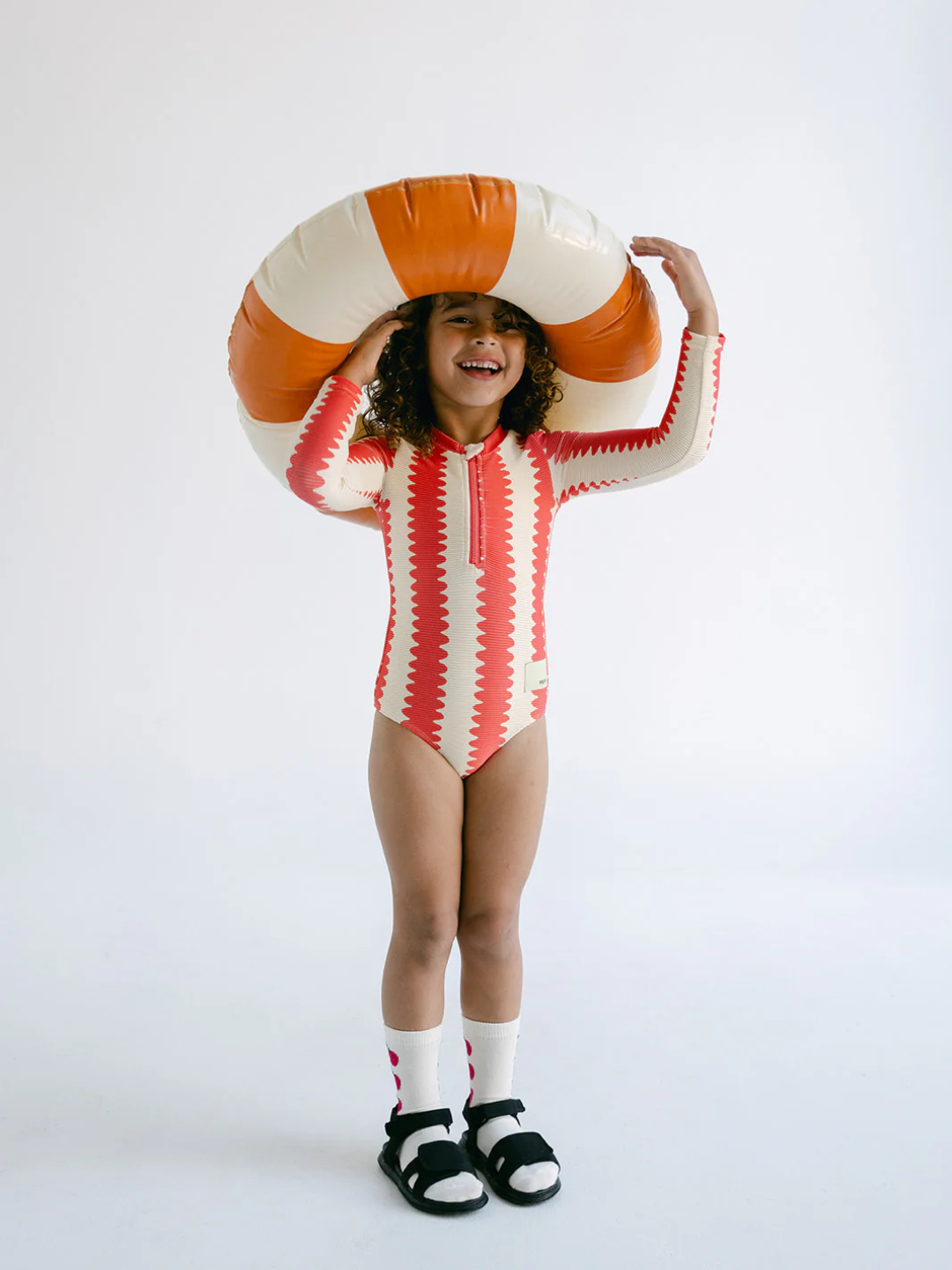 A child in a FULL PIECE SWIMSUIT made from recycled material smiles, holding an inflatable swim ring above their head against a plain backdrop.
