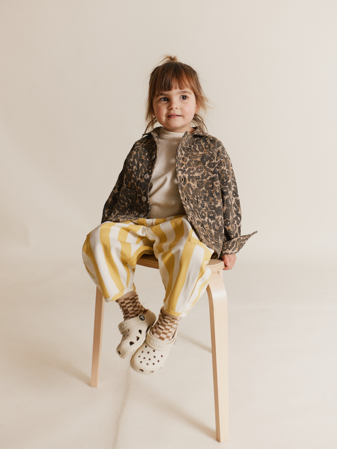 A young child sits on a wooden stool, wearing a leopard-print jacket, EMERSON HONEY STRIPE TROUSER, and white clogs.