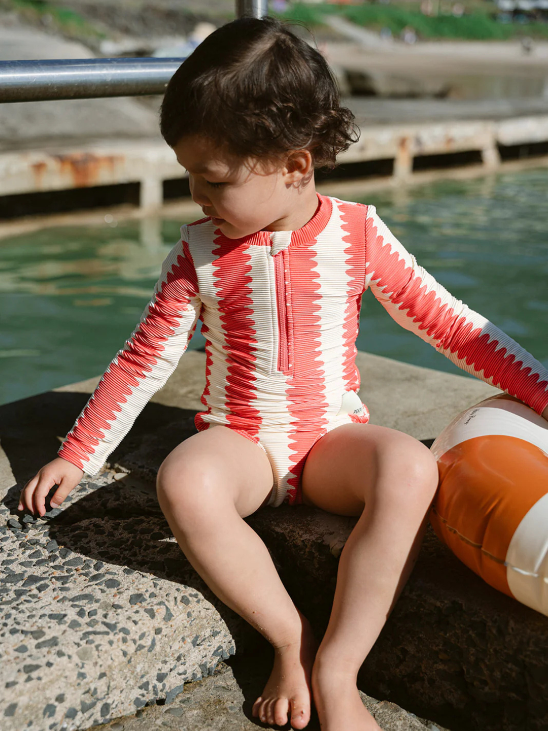 A young child in the FULL PIECE SWIMSUIT, a red and white long-sleeve design with UPF 50+ UV protection, sits by the water on a stone surface next to a buoy.