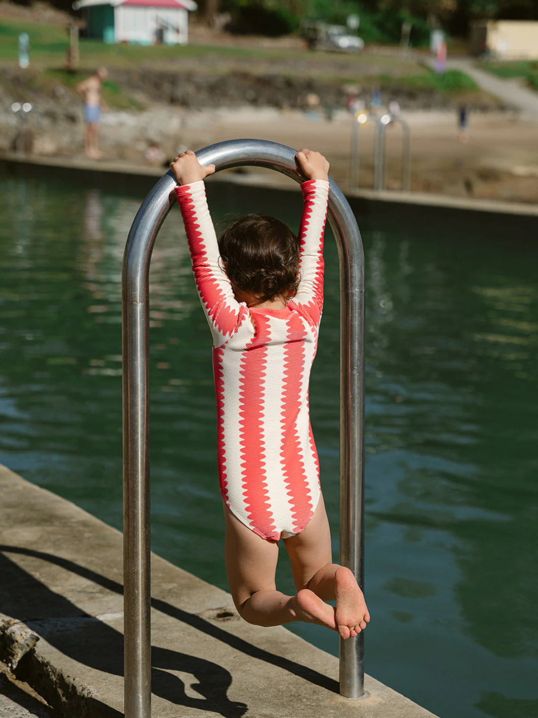 A child wearing a red and white striped FULL PIECE SWIMSUIT hangs from a metal ladder beside an outdoor pool, with water and a sandy shore in the background.