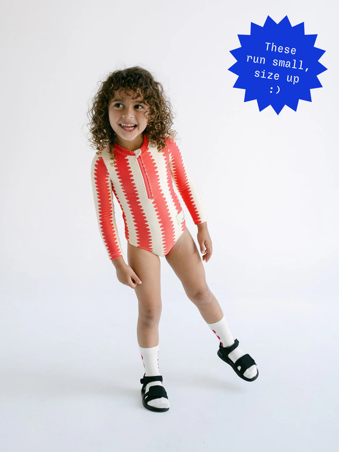A young child models the FULL PIECE SWIMSUIT, a red and beige patterned long-sleeve swimsuit made from recycled material, paired with white socks and black sandals. A blue label advises: "These run small, size up :).
