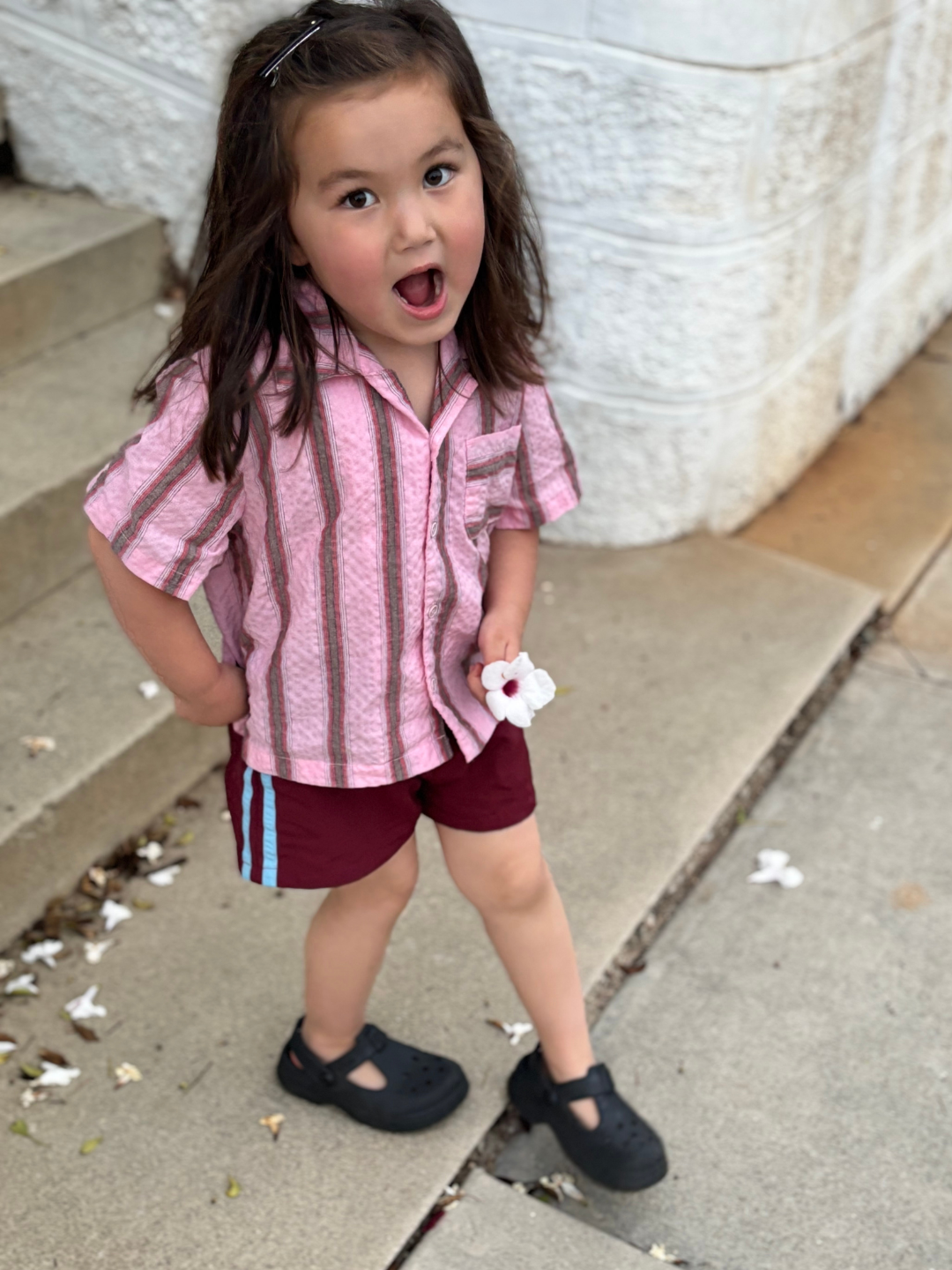 Maroon | A young child with long brown hair, wearing a pink striped shirt, black shoes, and SPRINTER SHORTS with an elastic waist, stands on a sidewalk holding a white flower and gazing upward with an open mouth.