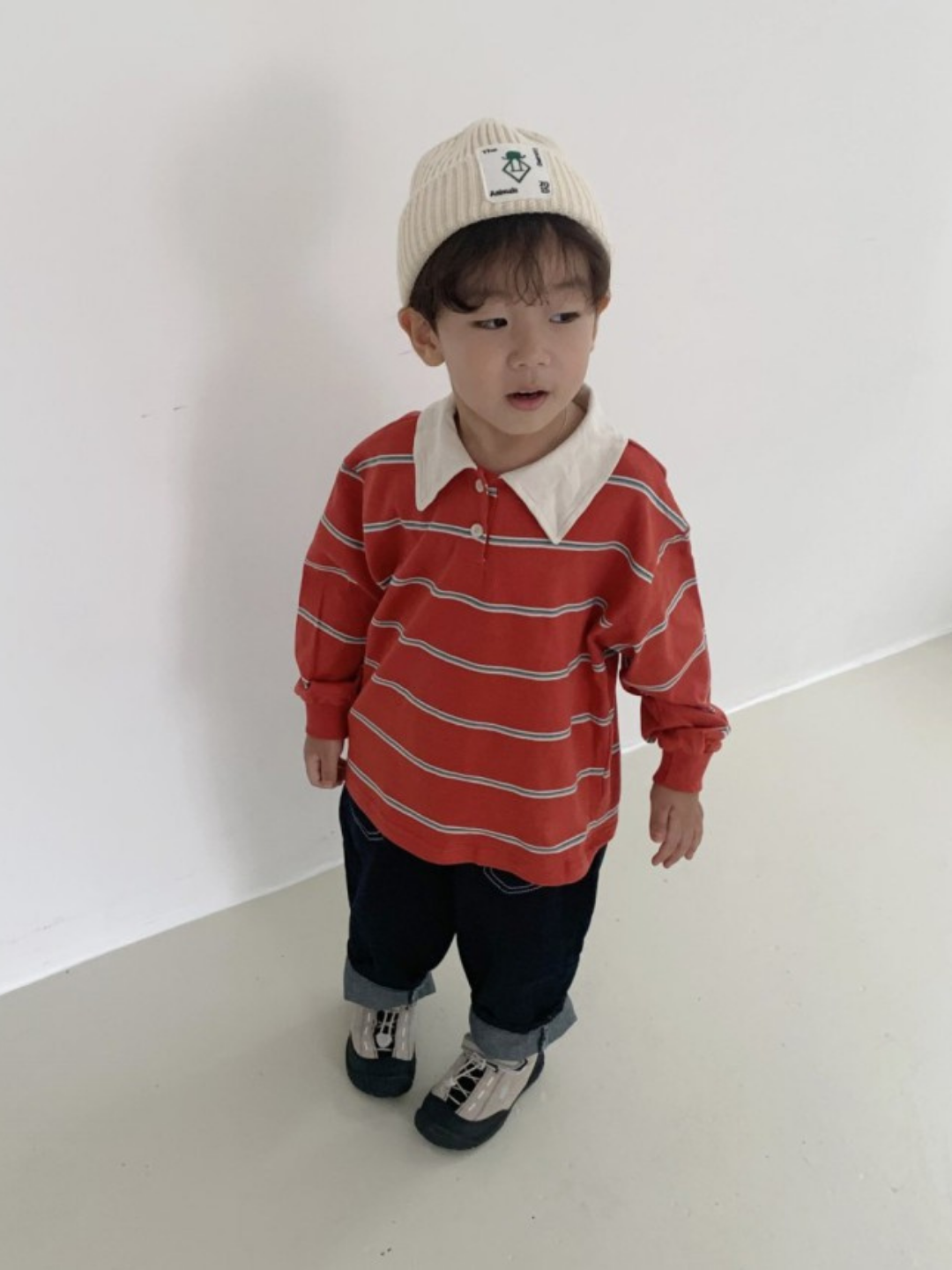 A young child wears a cream beanie, the RETRO RUGBY LONGSLEEVE (a Korean kids brand red striped tee with a rugby collar), dark pants, and sneakers, standing indoors against a white wall.