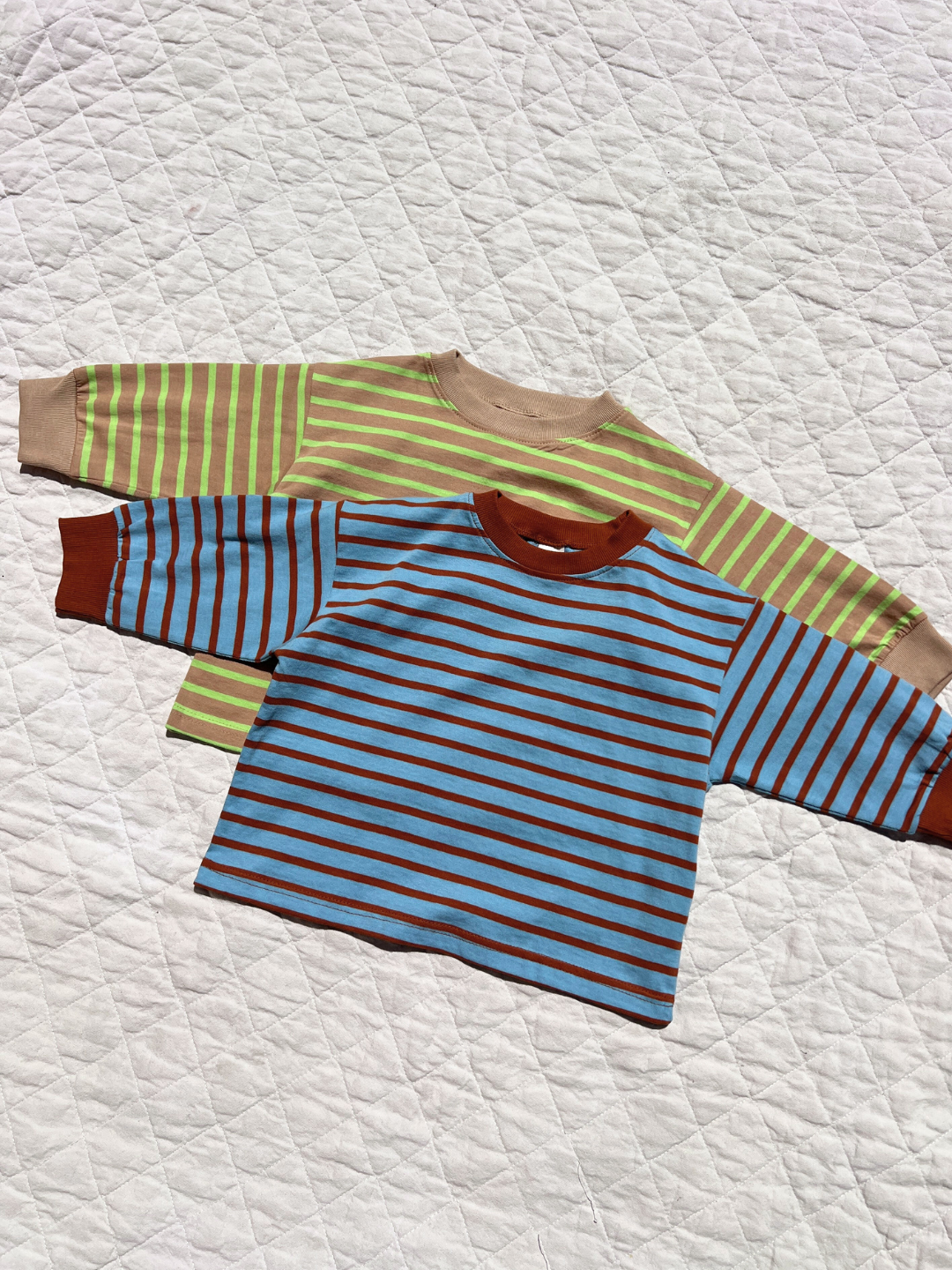 Two BAILEY STRIPE LONGSLEEVE tees from a Korean kids brand—one green with beige cuffs, one blue with brown stripes—are laid on a white quilted surface. Both are crafted from soft cotton for all-day comfort.