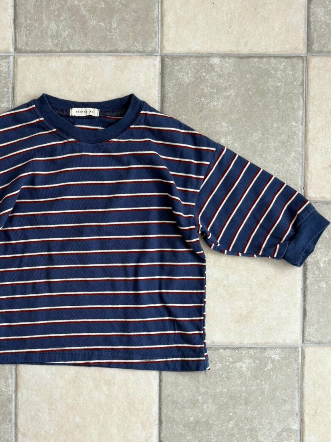 Blue | The PEPPER LONGSLEEVE TEE, a navy blue cotton top with thin white and red stripes from a Korean kids brand, is laid flat on a tiled floor.