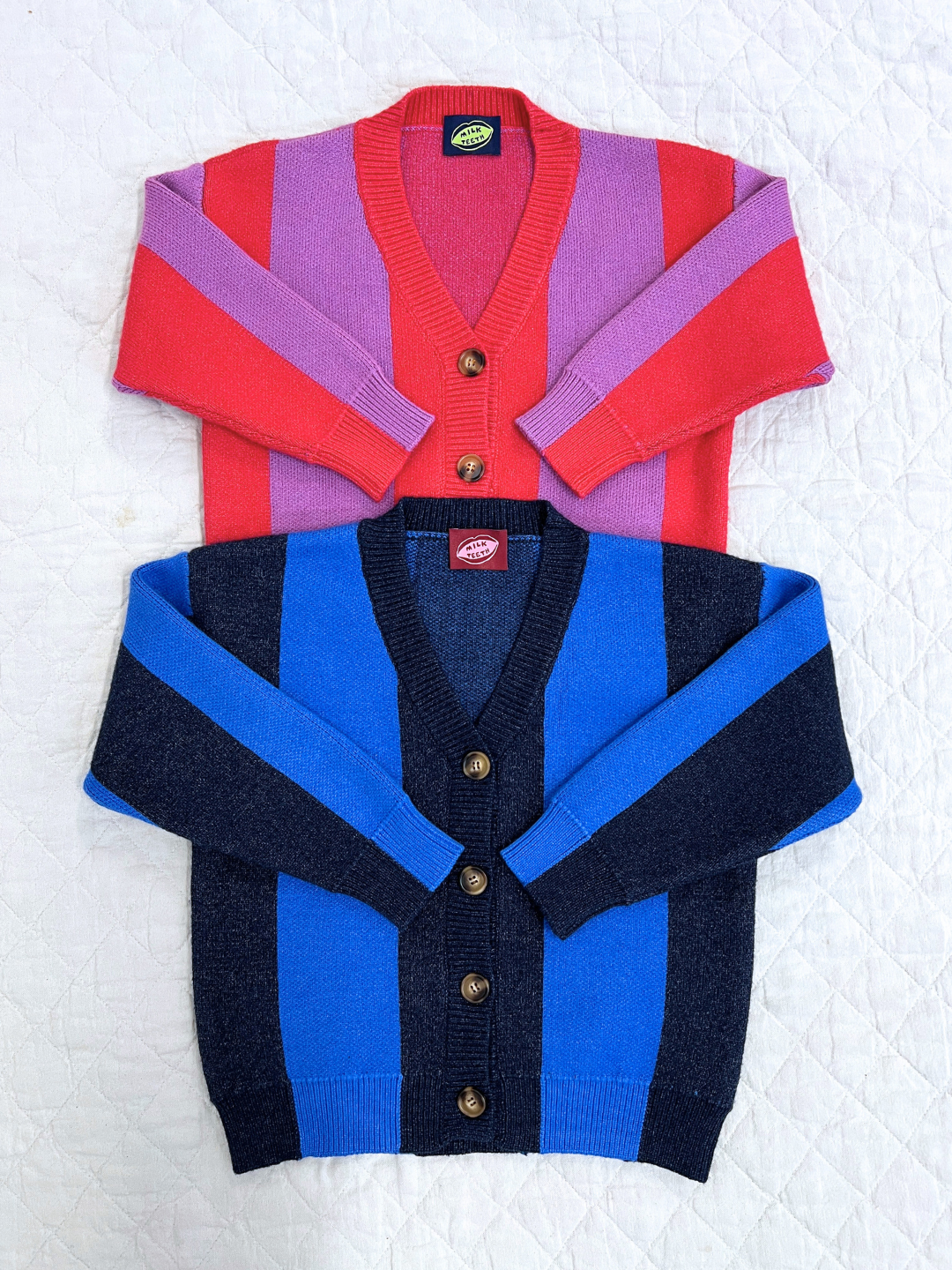 Blue | Two STRIPE CARDIGAN button-ups are laid flat on a white quilted surface: the top one features pink and red stripes, while the bottom one has blue and dark navy stripes.