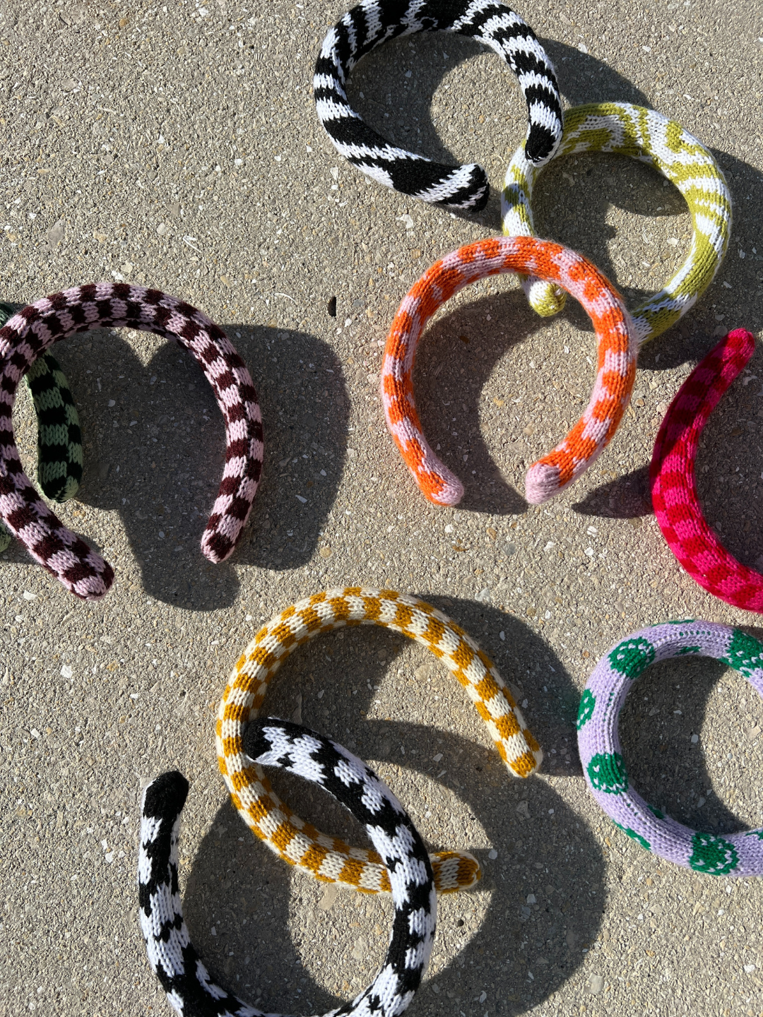 A collection of PUFFY KNIT HEADBANDs is spread out on a concrete surface, showcasing their vibrant, handmade designs. These unique headbands feature a variety of color combinations such as black and white, pink and brown, and orange and green. Some are adorned with stripes while others display checkered patterns.