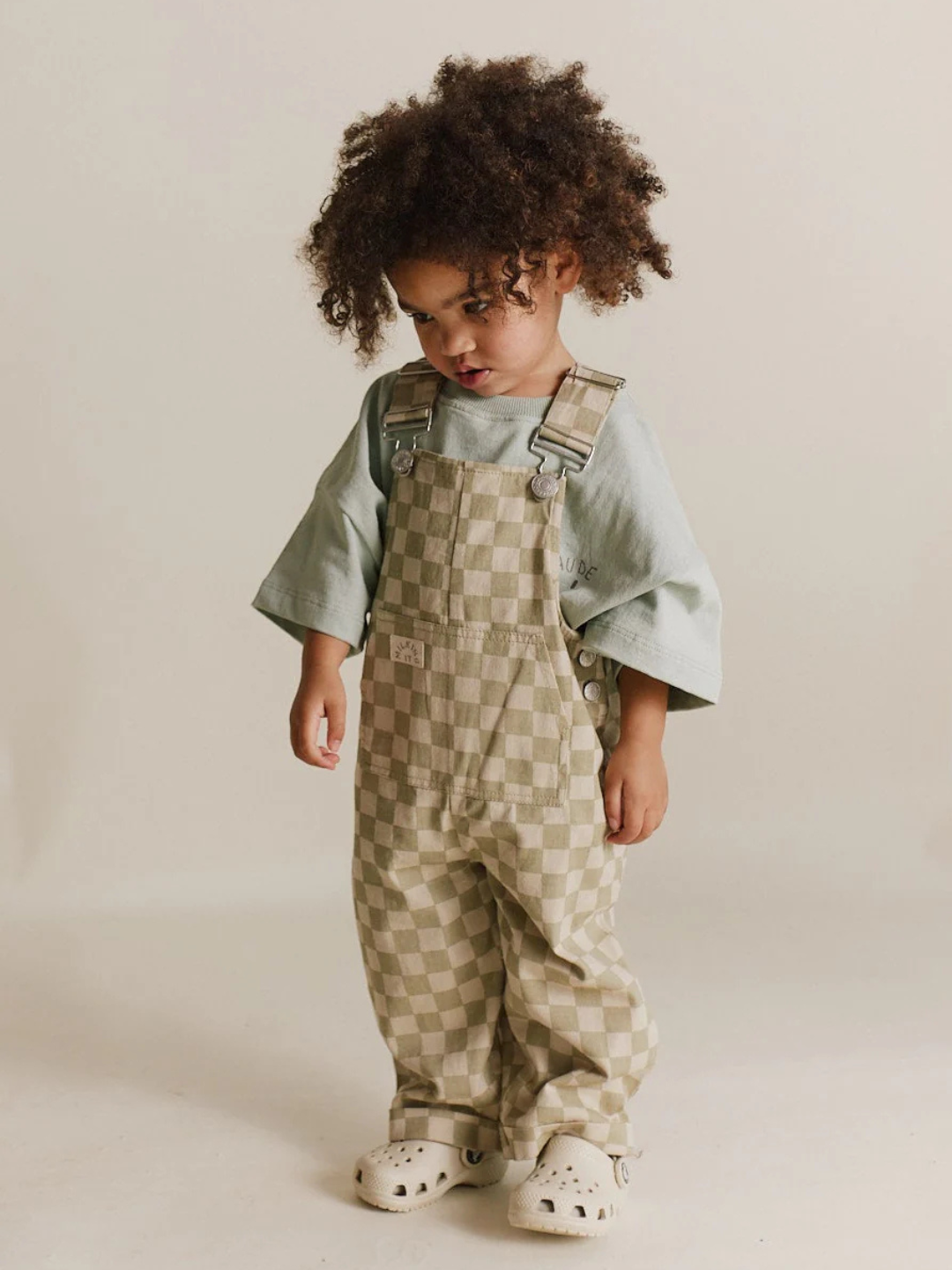 A curly-haired child stands wearing Claude & Co.'s Cecile Olive Check Dungaree, made from organic cotton twill, paired with a green shirt and beige sandals against a plain background.