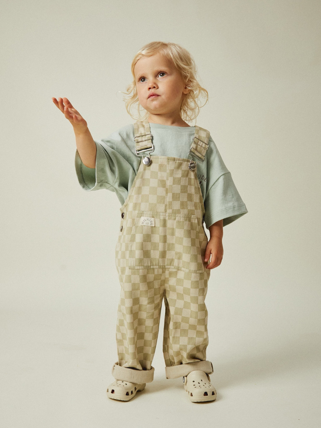 A blonde toddler stands against a neutral backdrop wearing Claude & Co.'s CECILE OLIVE CHECK DUNGAREE in charming checkerboard print, paired with a light green shirt and light-colored shoes. Made from soft organic cotton twill for all-day comfort, the toddler gestures upward with one hand.