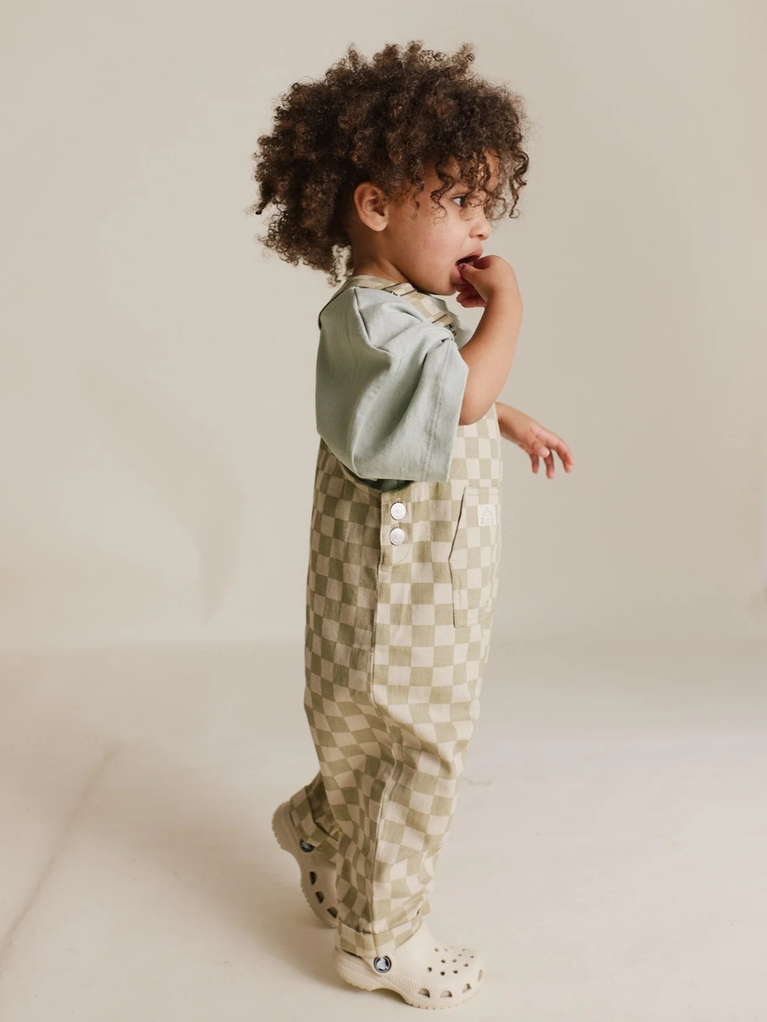 A curly-haired child stands sideways wearing Claude & Co's CECILE OLIVE CHECK DUNGAREES—crafted from soft organic cotton twill with buttons. The child has one hand near their mouth and is wearing clogs.