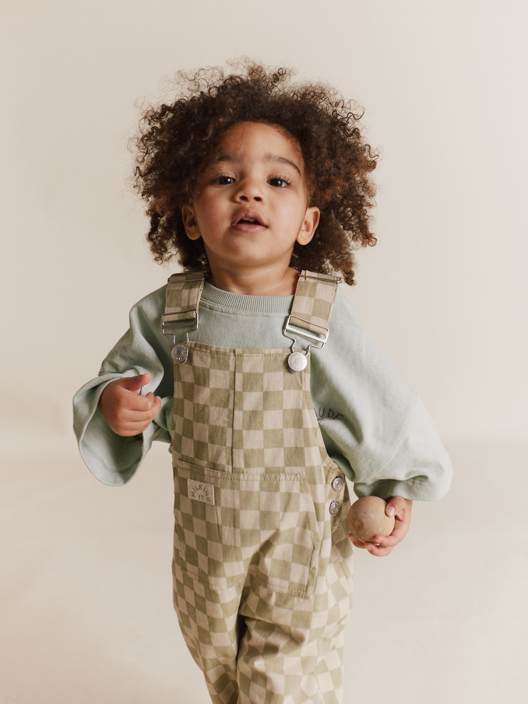 A young child with curly hair in a green sweater and Cecile Olive Check dungaree holds a wooden egg, standing against a neutral background.