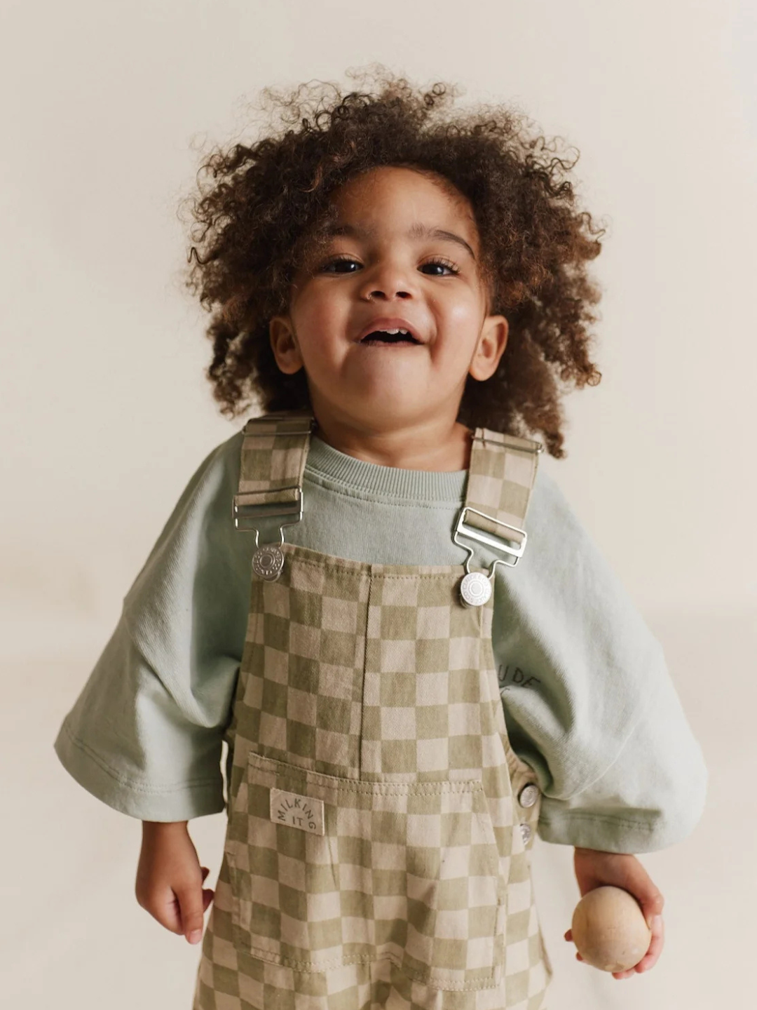 A curly-haired child beams in a green shirt and Claude & Co.'s Cecile Olive Check Dungaree, holding a small ball against a neutral backdrop.