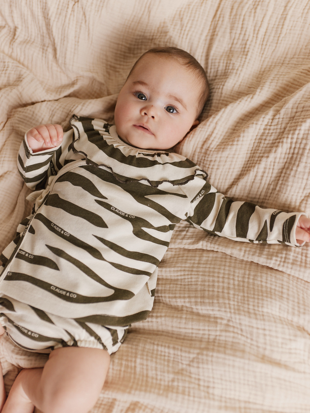 A baby lies on a beige bedspread, wrapped in the REMY ZEBRA WRAP CARDI SHORT SET made of organic cotton with olive stripes, looking upward with one arm outstretched.