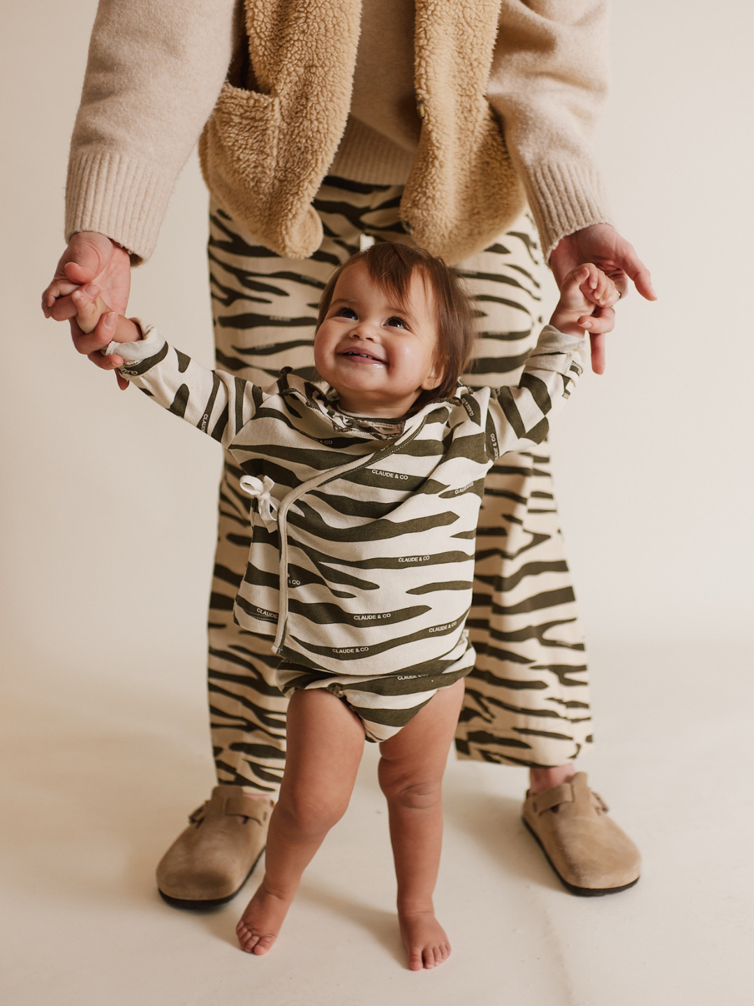 A toddler adorned in the REMY ZEBRA WRAP CARDI SHORT SET holds hands with an adult in matching pants and sweater, both beaming against a light backdrop. Their cozy organic cotton outfits make an ideal baby gift.