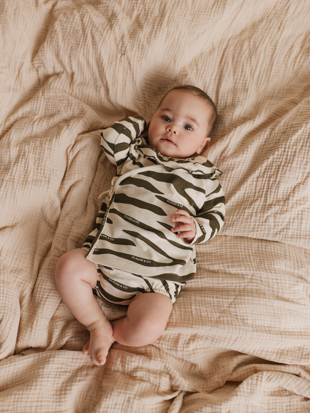 A baby, clad in the delightful REMY ZEBRA WRAP CARDI SHORT SET made from organic cotton, rests on a beige blanket, eyes on the camera with one arm raised—an adorable and thoughtful baby gift idea.