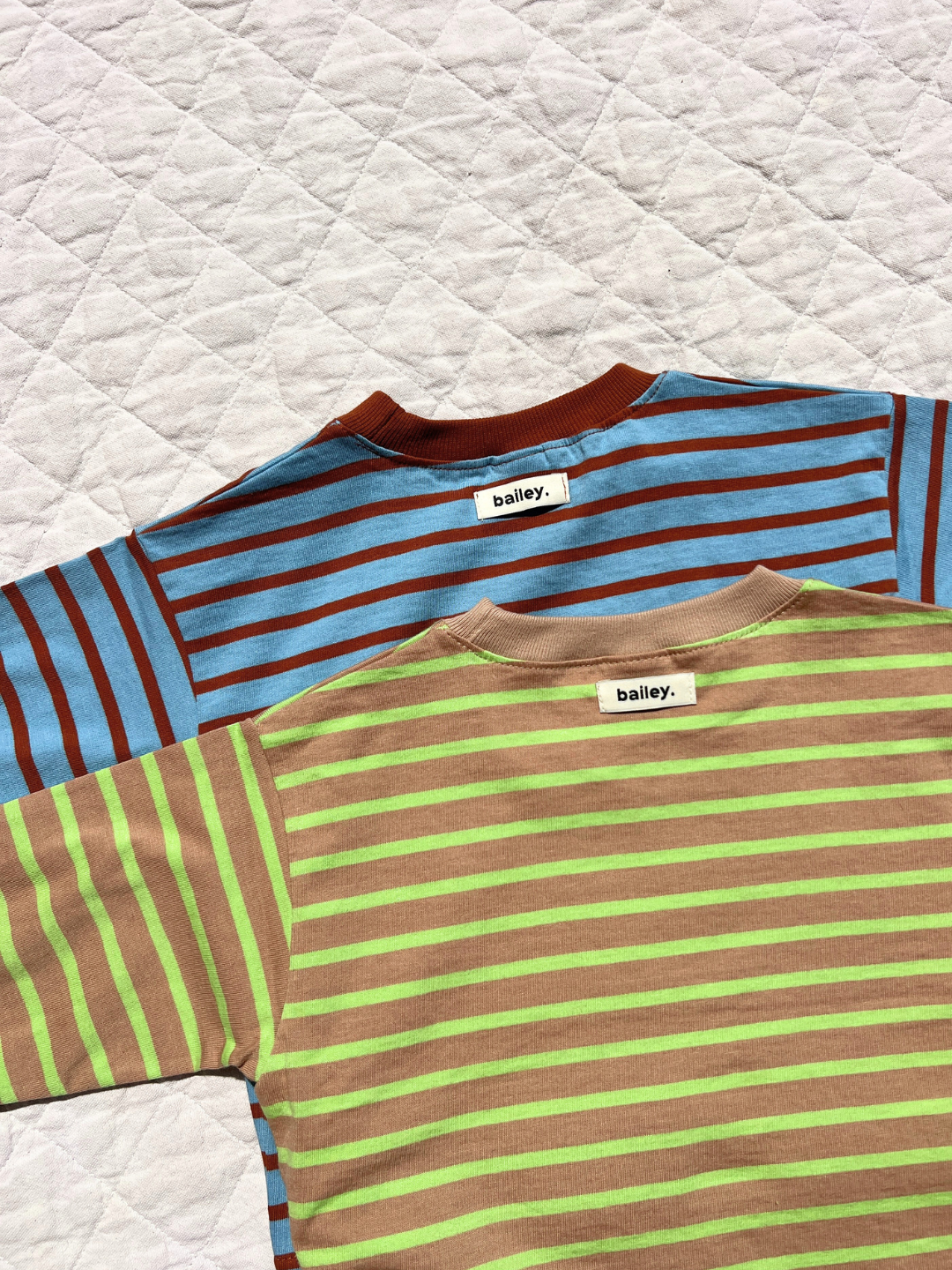 Two BAILEY STRIPE LONGSLEEVE tees from Korean brand "bailey."—one blue with brown stripes, the other beige with green stripes—made of soft cotton, are laid flat on a white quilted surface.