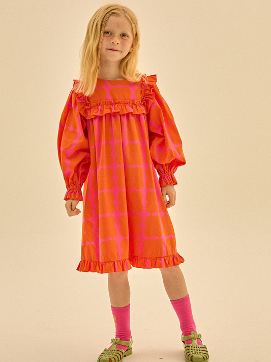 Second image of Front of Clover Dress. Bright red and pink dress with a ruffled hem, long sleeves with pleats, and ruffled trim on the collar.