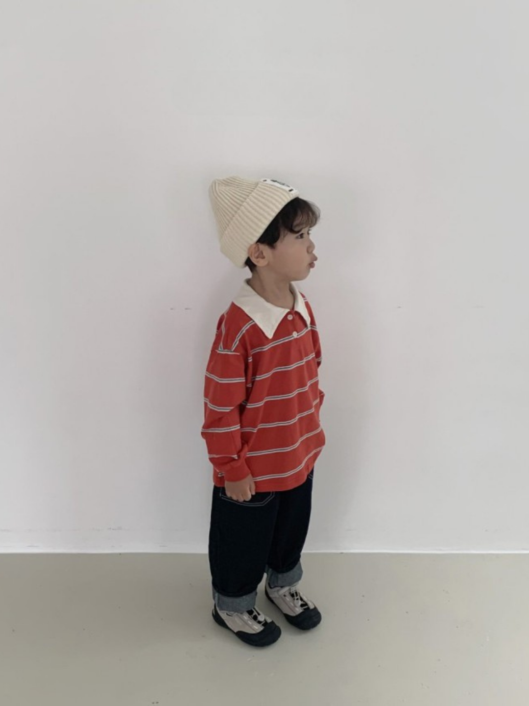 A young child stands indoors against a plain white wall, wearing the RETRO RUGBY LONGSLEEVE from a Korean kids brand, dark pants, a white beanie, and sneakers while looking to the side.