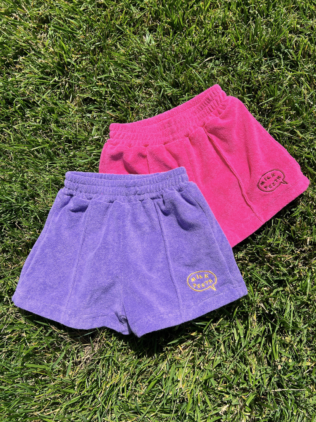 Two pairs of EVERYDAY TERRY SHORTS, one purple and one pink with yellow "MUK PEACH" logos and elasticated waistbands, are lying on green grass.
