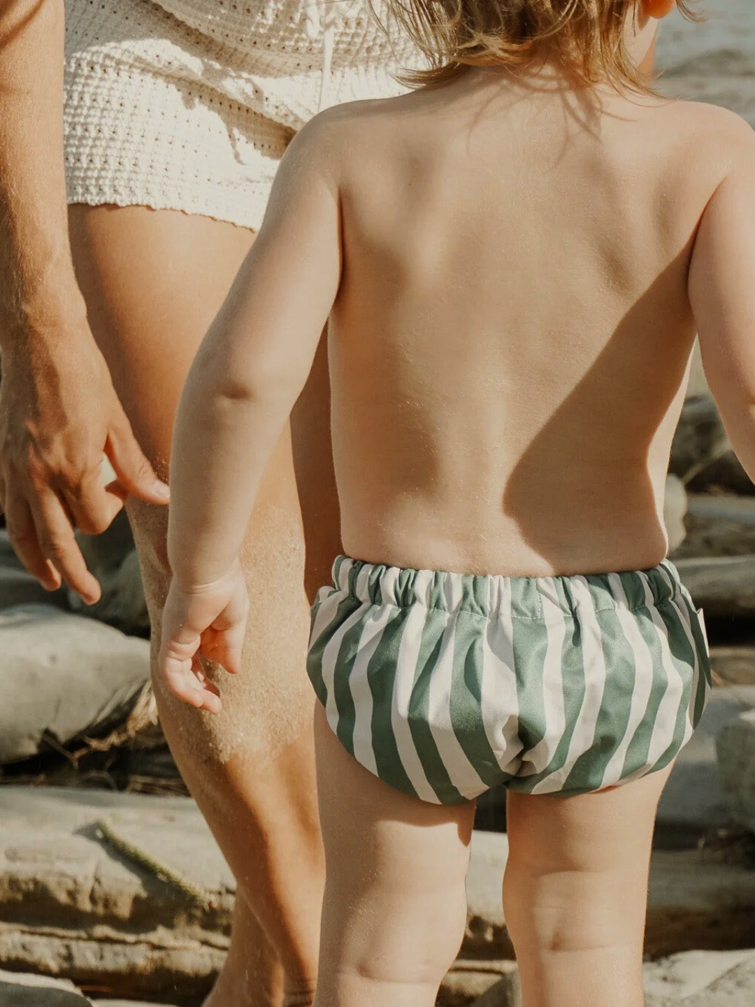 Jungle | A small child stands on a sandy beach in green and white striped SWIM DIAPER shorts made from buttery-soft recycled fabric.