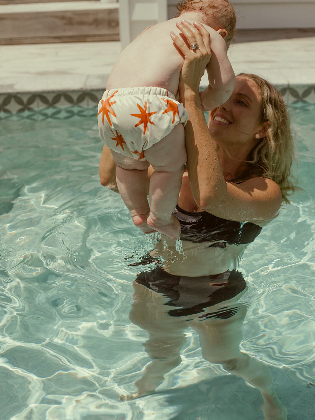 Capri | In a black swimsuit, a woman effortlessly lifts a baby in a SWIM DIAPER with orange star patterns as they enjoy the cool pool water.