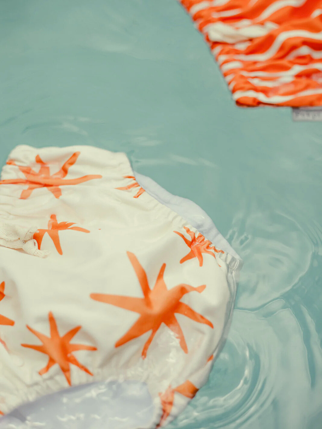 Capri | Two SWIM DIAPERs from Pekpi float: one in buttery-soft recycled fabric with orange starfish on white, the other showcasing an orange and white wave pattern.