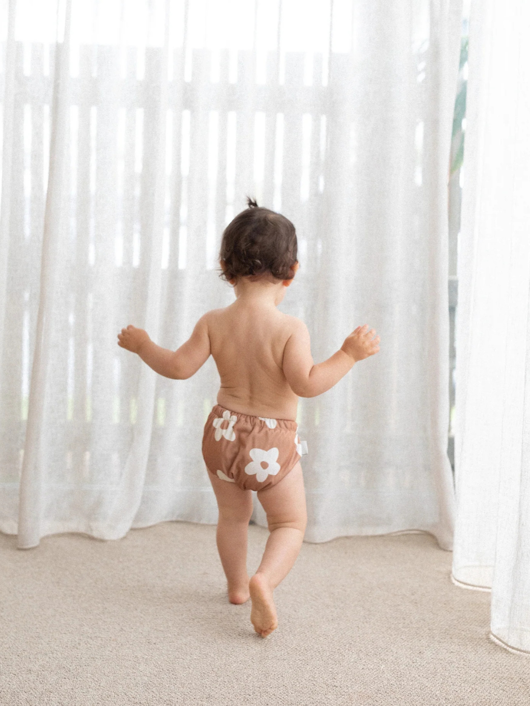 Posy | A toddler sporting floral-printed, buttery-soft recycled fabric SWIM DIAPER wobbles toward sheer white curtains in a softly lit room.