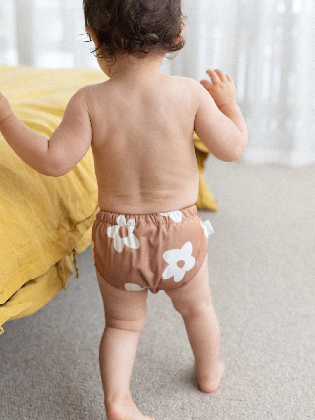 Posy | A toddler in SWIM DIAPER floral-patterned shorts, made from buttery-soft recycled fabric, stands beside a yellow bed facing away in a well-lit room.