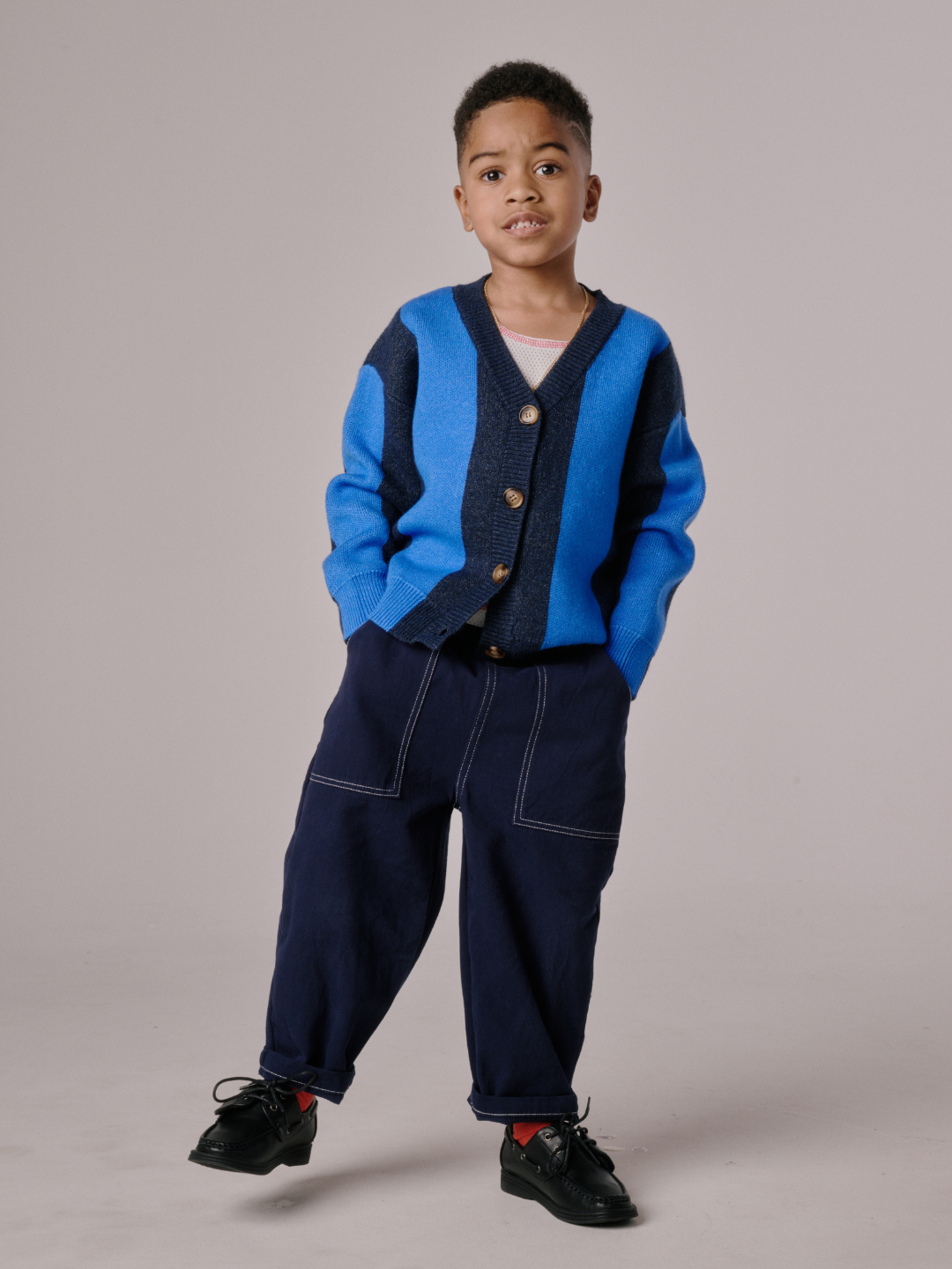 Blue | A young boy stands with his hands in his pockets, wearing the STRIPE CARDIGAN, dark pants, and black shoes against a plain background.