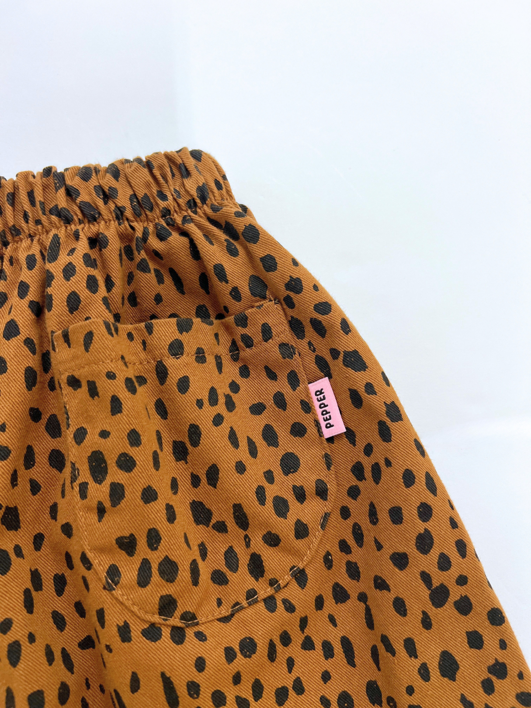 Brown | Close-up of MINI CHEETAH SPOT PANTS—brown cotton twill with black spots, elastic waistband, pocket, and a pink "PEAR" tag—a playful detail from a unisex kids brand.