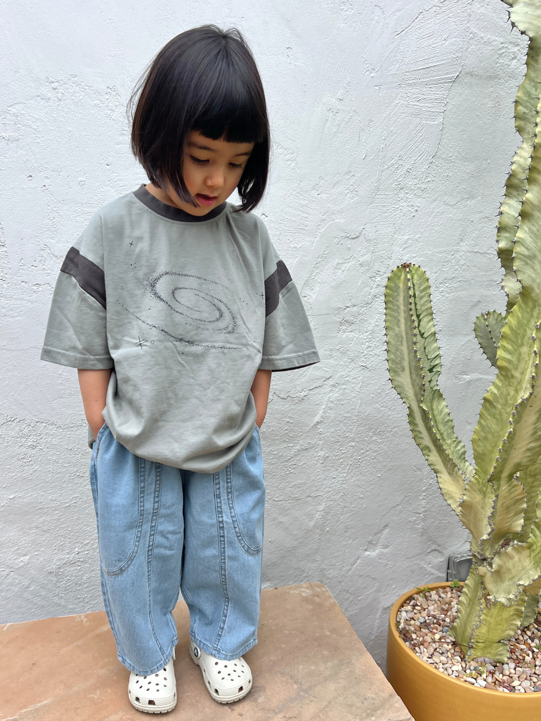 A young child with short dark hair stands next to a tall potted cactus, wearing a loose gray swirl shirt, MIYU JEANS—lightweight baggy denim with an elasticated waist—and white shoes.