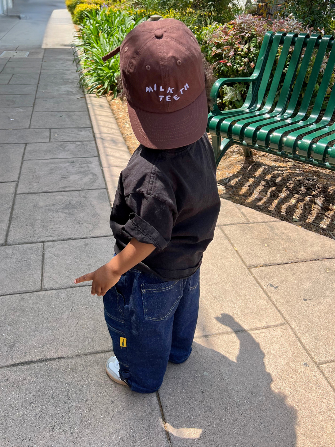 Brown | Child wearing the Milk Teeth baseball cap in brown, worn backwards. He is wearing a black t-shirt and baggy dark blue jeans, and standing on a sidewalk in front of a green bench.