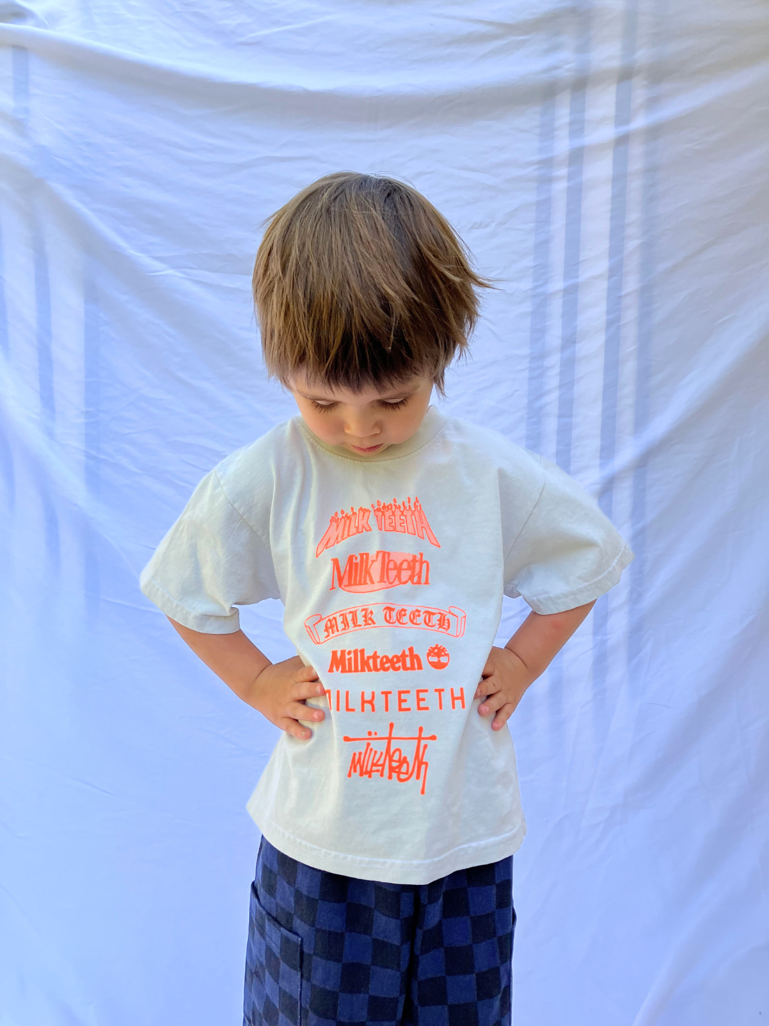 Cement | A child wearing a light grey t-shirt with Milk Teeth spelled out in various fonts in orange ink. He wears navy and black checkered pants and is standing in front of a white sheet backdrop.