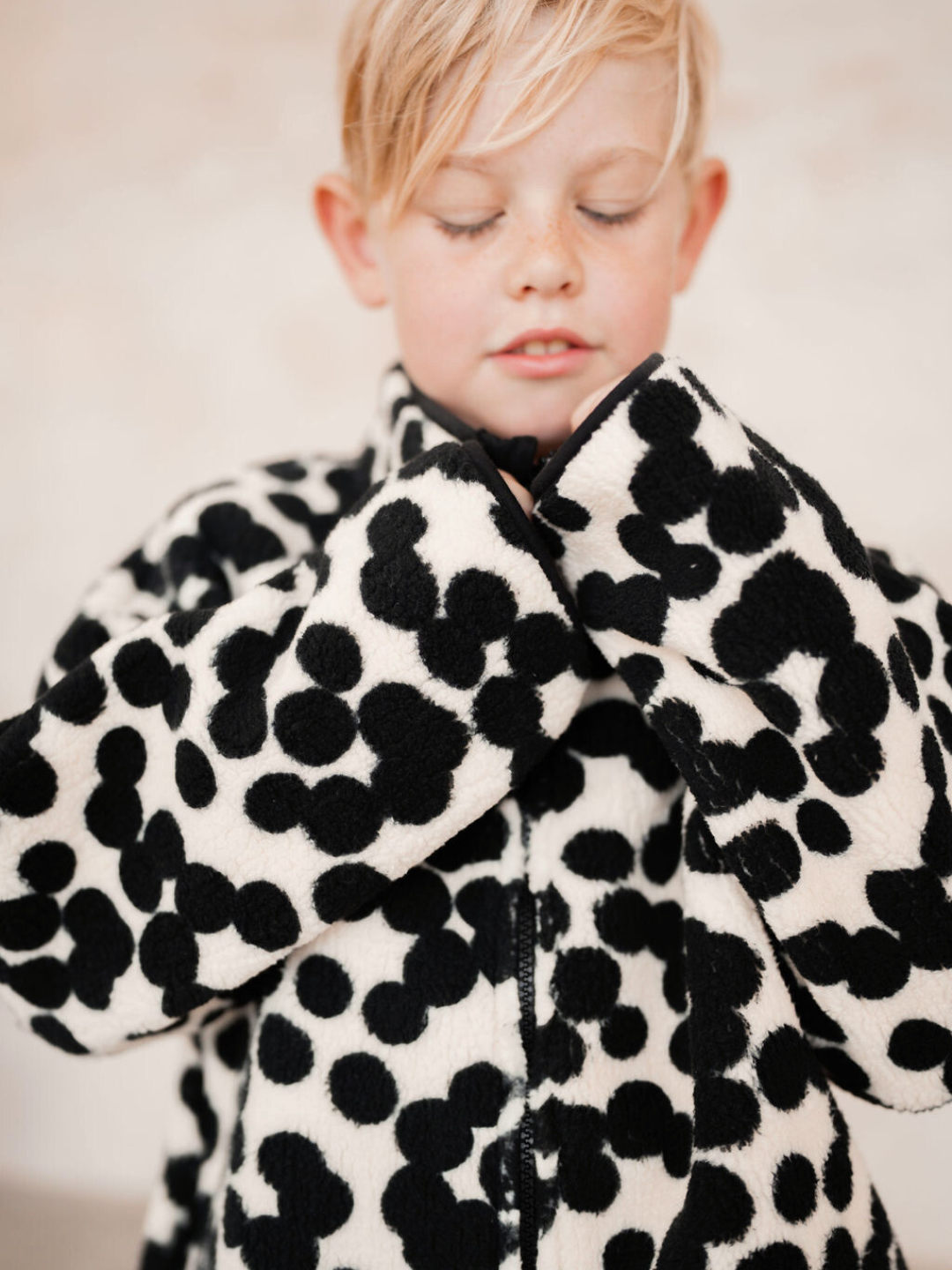 A child with closed eyes is wearing the SPOT FUNNEL COAT, a black and white polka-dotted jacket. Shop all clothing to discover similar adorable styles.