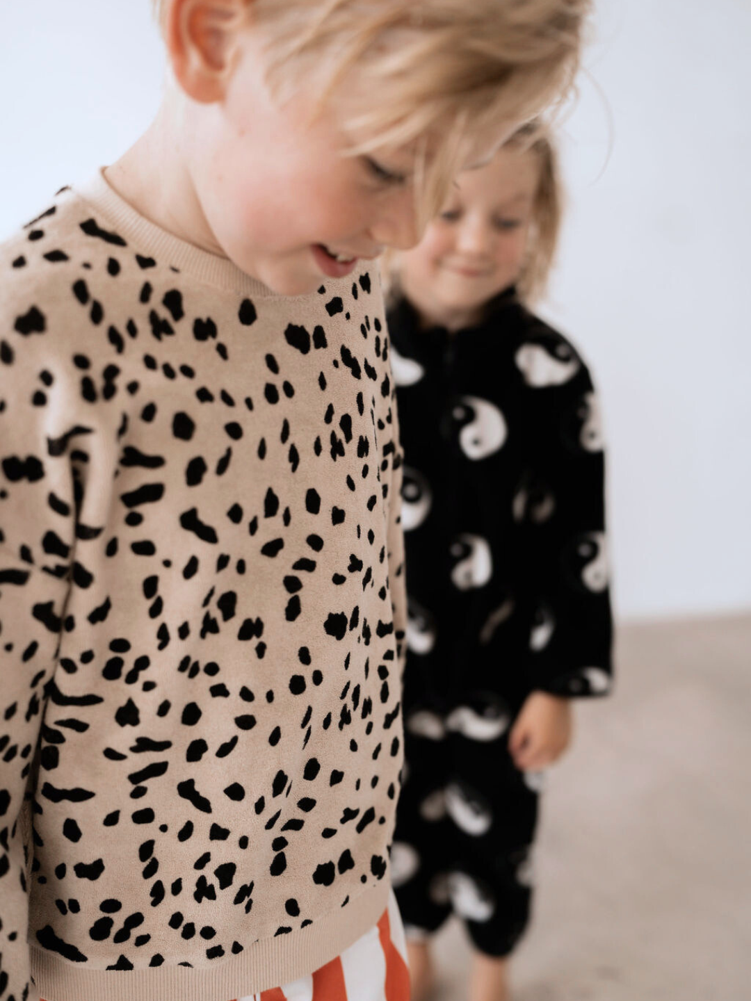 Two children are standing indoors. The baby in the foreground wears a DALMATIAN TERRY SWEATSHIRT and red striped pants. The child in the background wears a black outfit with white patterns.