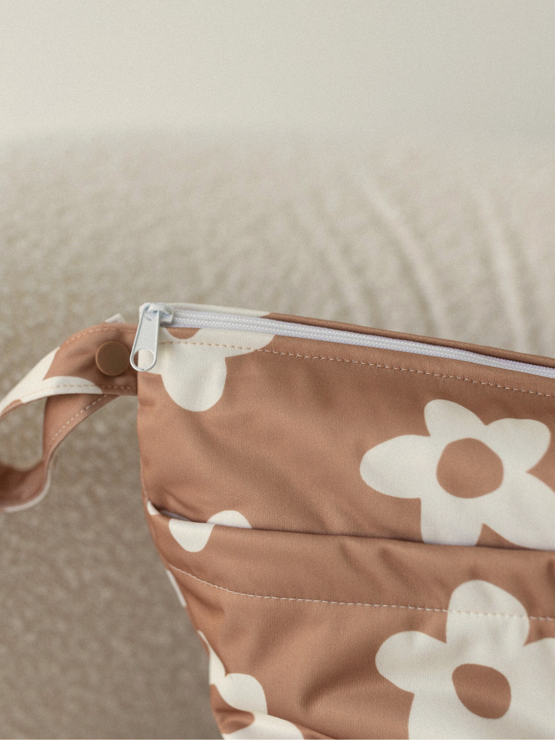 Posy | A close-up of the WET BAG, featuring brown fabric with a white floral pattern and a white zipper, resting on a textured light surface—ideal for carrying swim diapers or essentials.