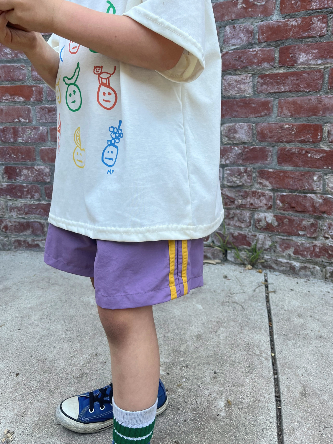 Purple | A child stands on concrete, wearing a white shirt with colorful faces, SPRINTER SHORTS with an elasticated waist, green socks, and blue sneakers. A brick wall is in the background.