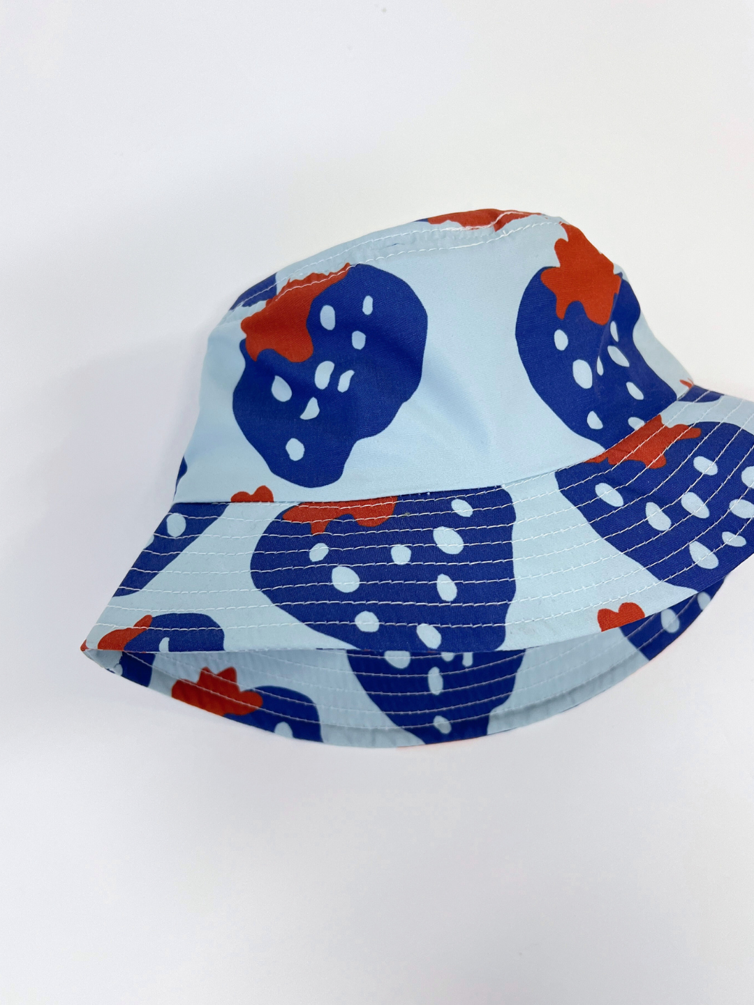 The STRAWBERRY BUCKET HAT features a blue strawberry print with red highlights and white dots on a light blue background, made from sustainable materials. Positioned gracefully on a white surface, it captures the vibrant essence of Seaetsa Surf.