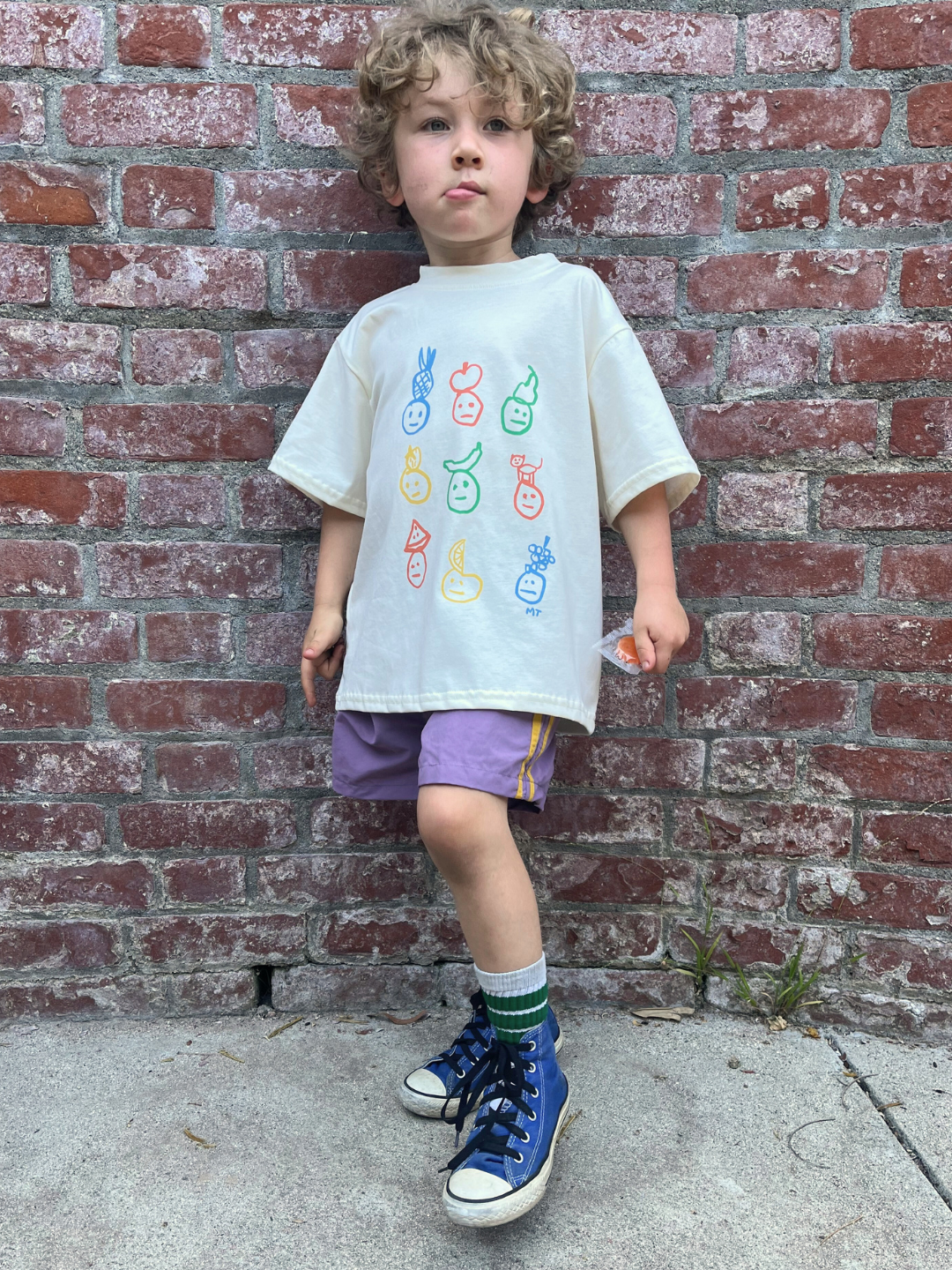 Purple | A young child with curly hair stands by a brick wall, wearing SPRINTER SHORTS with an elastic waist, a white t-shirt with colorful fruit faces, striped socks, and blue high-top sneakers.