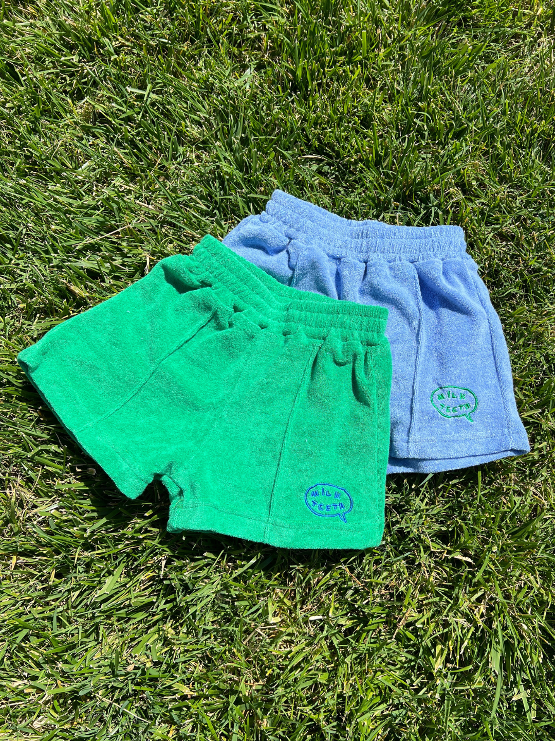 Two pairs of EVERYDAY TERRY SHORTS for children, one green and one blue with elasticated waists, are side by side on grass in bright sunlight.