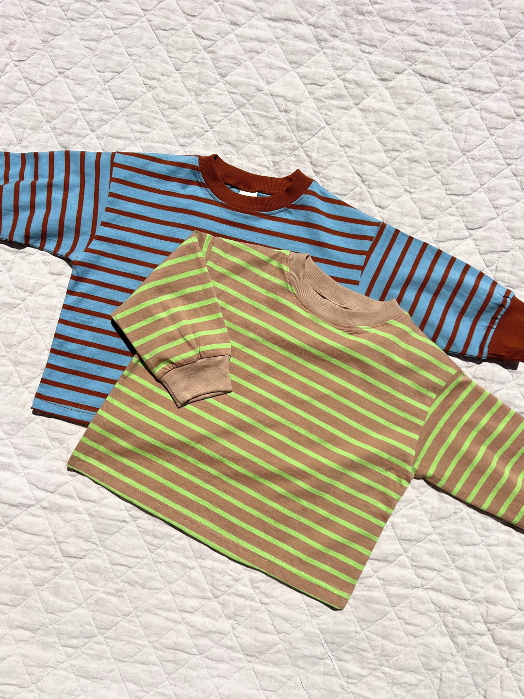 Two BAILEY STRIPE LONGSLEEVE tees from a Korean kids brand in soft cotton—one blue and brown, the other green and beige—are laid out on a white quilted surface.