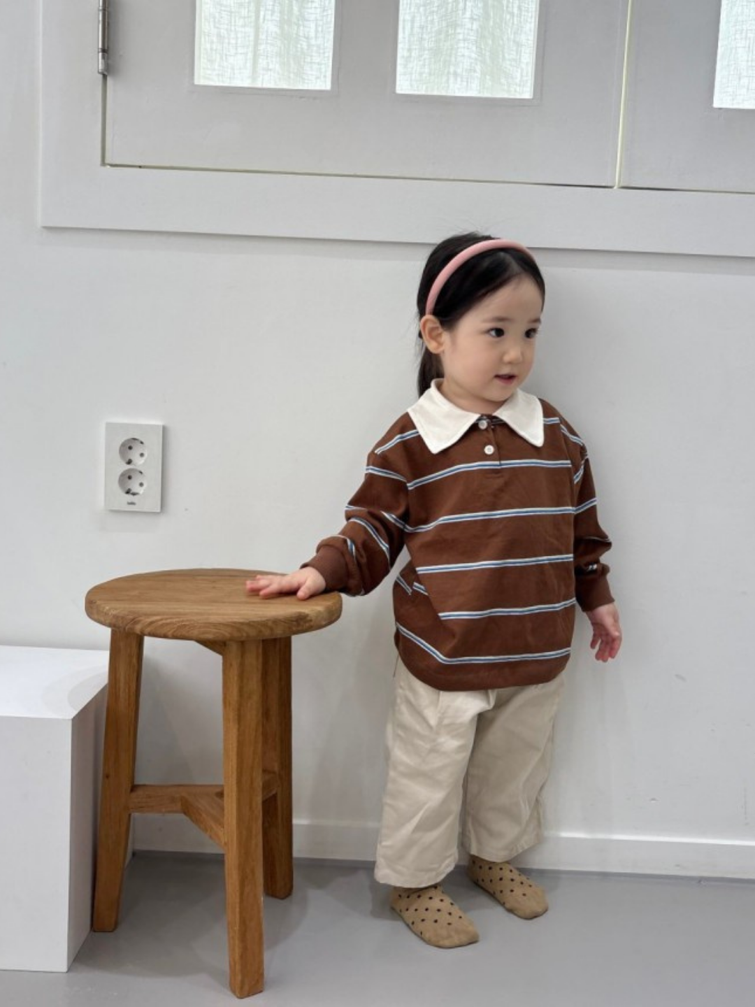 A young child in the RETRO RUGBY LONGSLEEVE, cream pants, and a pink headband stands by a wooden stool in a minimal room, perfectly capturing the cozy vibe of a Korean kids brand.