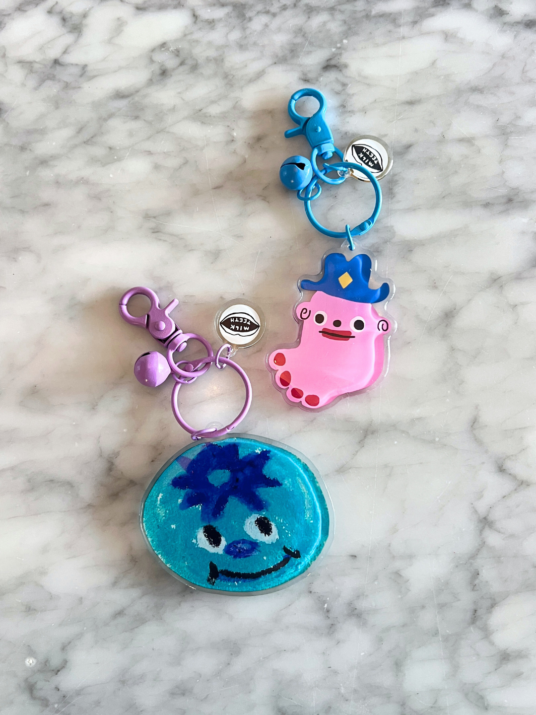 Blueberry | SOUVENIR KEYCHAIN by Katie Kimmel: two character charms—a pink one with a crown and a blue round one with a smiling face—feature clips and small round tags, displayed on a marble surface as playful bag accessories.