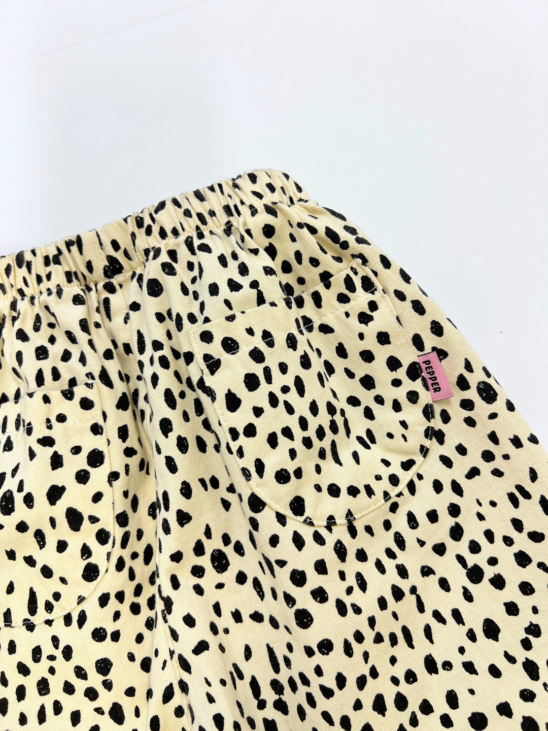 Off-White | Close-up of MINI CHEETAH SPOT PANTS—cream-colored cotton twill with black cheetah spots, two back pockets, and a pink label on the right pocket from a unisex kids brand.