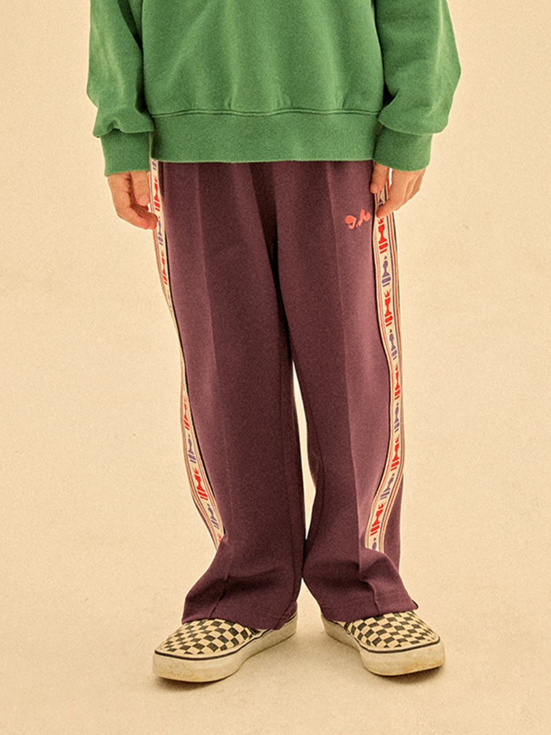 Purple | A person in a green sweatshirt, CHESS TRACK PANTS with purple side stripes, and black-and-white checkered slip-on shoes stands on a light surface