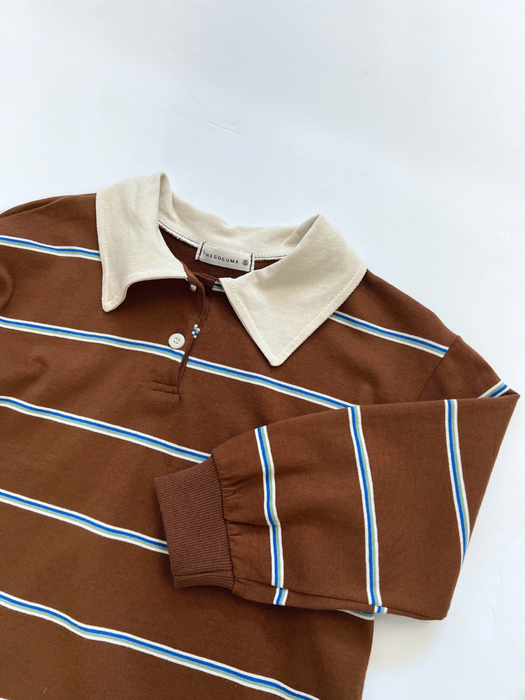 The RETRO RUGBY LONGSLEEVE from a Korean kids brand features blue and white stripes, a beige rugby collar, brown buttons, and is shown laid flat on a white surface.