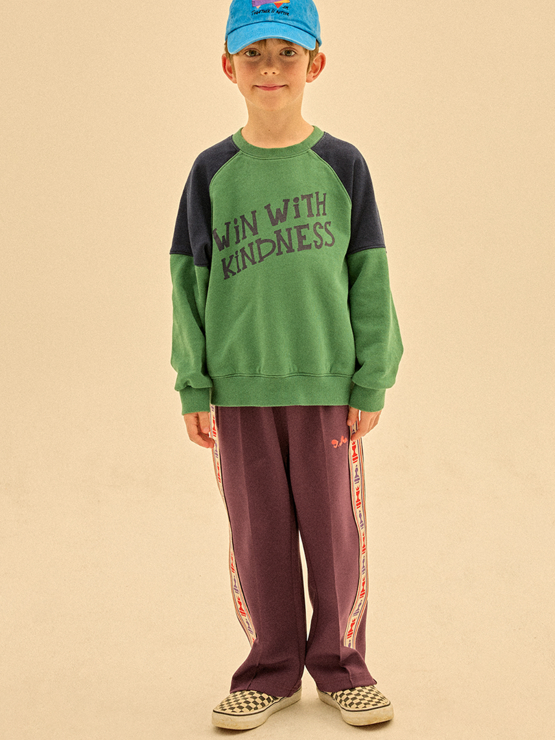 Purple | A young child in a blue cap, green "Win With Kindness" sweatshirt, CHESS TRACK PANTS with side stripes, and checkered slip-on shoes stands facing the camera.