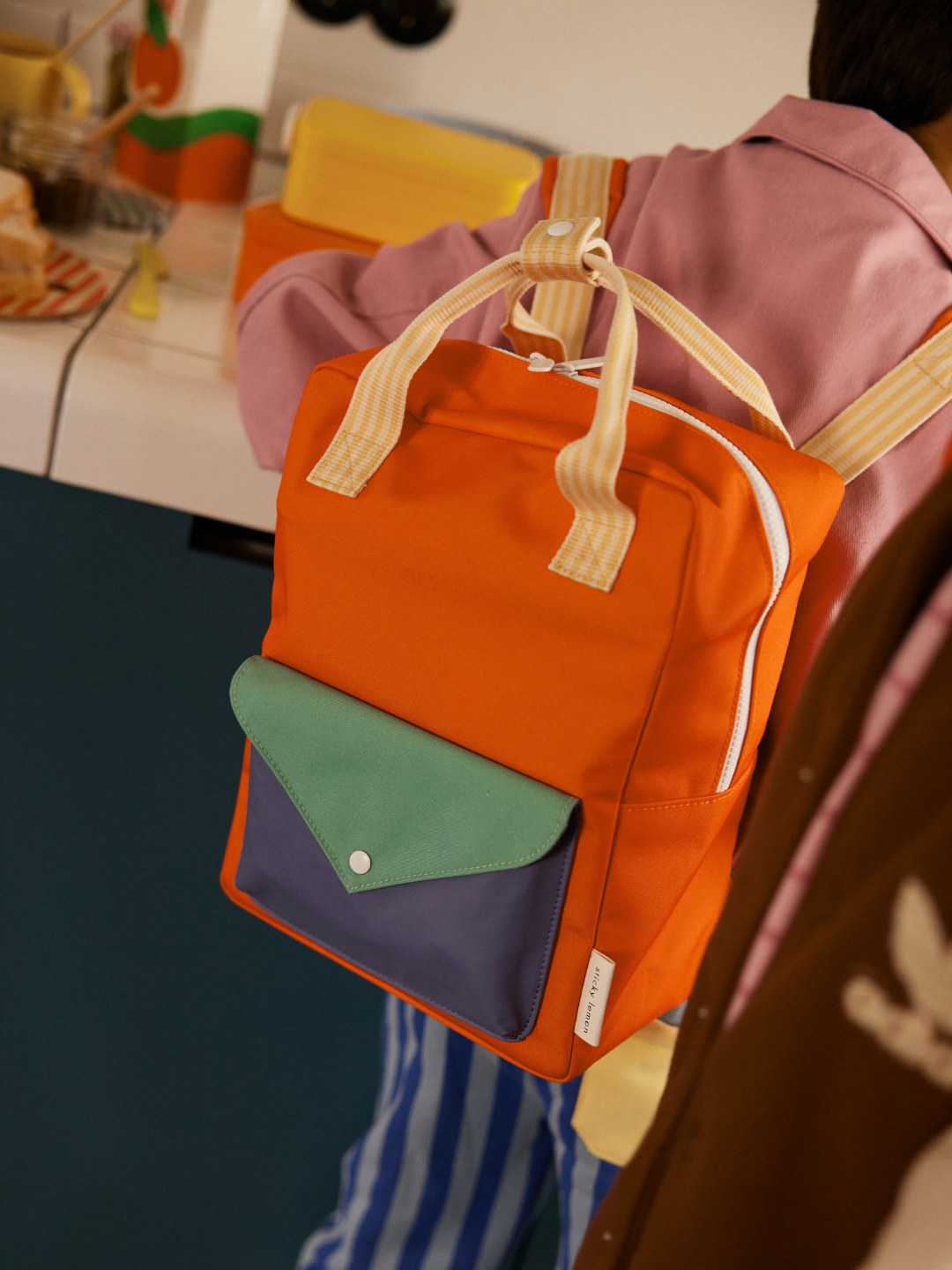 Lifebuoy Orange | A person in a pink shirt and blue striped pants carries the MEDIUM ENVELOPE BACKPACK—orange, medium-sized, with a green and purple front pocket, crafted from recycled materials.